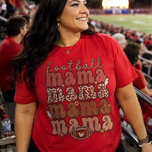 以下が含まれることがあります： 4段にわたって「football mama」というテキストが繰り返された赤い半袖Tシャツ。デザインには、ヒョウ柄、無地の茶色、輪郭線で描かれた文字が含まれており、小さな星、稲妻、フットボールのアイコンがアクセントとしてあしらわれています。