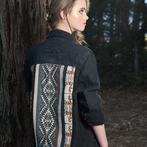 Könnte beinhalten: Schwarze Jeansjacke mit dekorativem Rückenteil. Das Panel zeigt ein geometrisches Design in Weiß-, Grau-, Braun- und Blautönen. Die Jacke hat lange Ärmel und einen klassischen Kragen. Das Model steht in einem Wald.