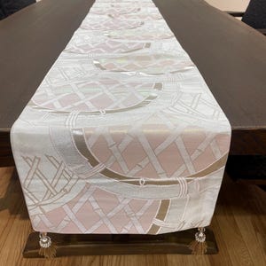 以下が含まれることがあります： 白、ピンク、ゴールドの色調で繰り返される幾何学模様の長方形のテーブルランナー。 ランナーは濃い茶色の木製テーブルの上に置かれています。 装飾的なタッセルが端からぶら下がっています。