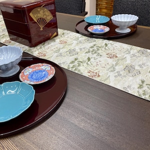 以下が含まれることがあります： 金色の扇のデザインが施された、漆塗りの多層弁当箱と、さまざまなサービング皿が特徴のテーブルセッティング。 ターコイズ色のプレート、小さな模様入りのプレート、白いボウルが濃い木製のトレイに載っています。模様入りのテーブルランナーも存在します。