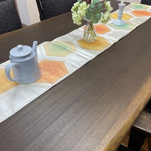 以下が含まれることがあります： 濃い茶色の長い木製ダイニングテーブルには、オレンジ、黄色、緑色の幾何学模様のテーブルランナーが敷かれています。水色のティーポット、花瓶に入った花、ろうそく立てとろうそくがテーブルに置かれています。
