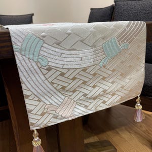 May include: A beige and white table runner with a woven pattern and decorative rope-like designs in light blue and pink. The runner is adorned with tassels at each end, adding a touch of elegance to the table setting.