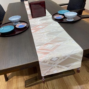 以下が含まれることがあります： 模様入りのテーブルランナー、赤い漆塗りの箱、いくつかのサービング皿がセットされた、濃い木製のダイニングテーブル。 テーブルランナーは、ベージュ、ピーチ、グレーのダイヤモンド柄です。 食器は青と白です。