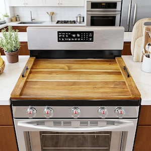 May include: A stainless steel gas range with a wooden stove cover. The cover has a rectangular shape with black handles. The range has five burners and a digital display. The kitchen setting includes a plant and cutting boards.