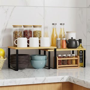 May include: A kitchen countertop arrangement featuring two-tiered wooden shelves with black metal supports. The shelves hold white mugs, glass jars with wooden lids, bowls, and spice racks. Various bottles and a black teapot are also present.
