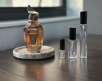 Op de afbeelding: Een amberkleurige parfumfles met een gouden dop en het woord "FALL" op de voorkant, op een grijze stenen onderzetter. Er staan ​​verschillende lege glazen parfumsprayflessen en een monsterflesje naast.