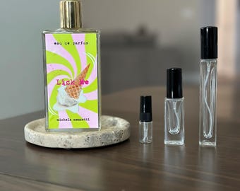 Op de afbeelding: Een rechthoekige glazen parfumfles met een gouden dop en een etiket met een roze en groen wervelpatroon, een ijshoorntje en de tekst "eau de parfum Lick Me michela mezzetti," naast drie kleinere doorzichtige glazen monsterflesjes.
