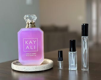 Op de afbeelding: Een grote rechthoekige parfumfles met een paarse iriserende afwerking, een diamantvormige dop en een etiket met de tekst KAY ALI VANILLA CANDY ROCK SUGAR | 42, vergezeld door drie kleinere doorzichtige glazen verstuivers met zwarte doppen.
