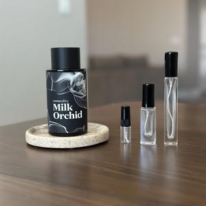 May include: A collection of perfume bottles and a small sample vial on a wooden surface. The largest bottle is black with the text "commodity Milk Orchid" and a floral design. The other bottles are clear glass with black caps.