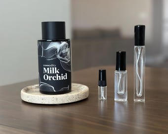 Op de afbeelding: Een verzameling parfumflesjes en een kleine proeffles op een houten oppervlak. De grootste fles is zwart met de tekst "commodity Milk Orchid" en een bloemenontwerp. De andere flessen zijn van helder glas met zwarte doppen.