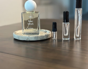 Op de afbeelding: Een doorzichtige glazen parfumfles met een witte bolvormige dop met het label Liis Rose Struck Eau de Parfum, rustend op een gespikkeld stenen dienblad, vergezeld door drie kleinere glazen verstuivers met zwarte doppen van verschillende hoogtes.