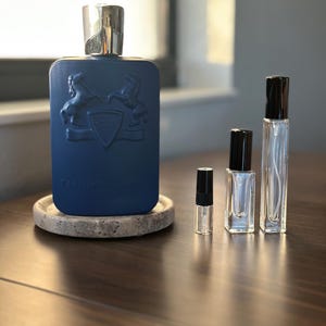 May include: A dark blue perfume bottle with a silver cap and embossed design, accompanied by three smaller sample bottles. The main bottle sits on a round, speckled coaster. The bottles are arranged on a dark wooden surface.