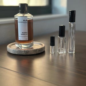 May include: A perfume bottle and several smaller vials are displayed on a wooden surface. The main bottle, labeled "Roses Vanille," is filled with a brown liquid. The smaller vials are clear with black caps.