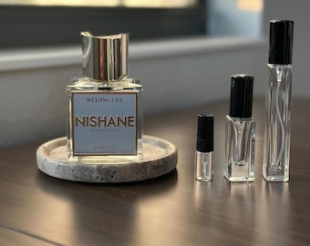 Op de afbeelding: Een parfumfles met een zilveren dop en een label met de tekst "Wulong Cha Nishane Shanghai Extrait de Parfum" staat op een ronde stenen tray. Verschillende kleinere, lege parfumflesjes zijn rechts gerangschikt.