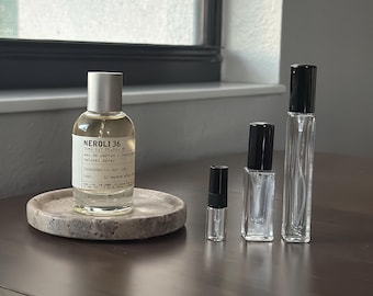 Op de afbeelding: Een verzameling parfumflessen en vials. Een grote fles met een zilveren dop staat op een ronde, gespikkelde stenen tray. De fles is gelabeld met "NEROLI 36". Verschillende kleinere, heldere glazen vials met zwarte doppen zijn in de buurt gerangschikt.