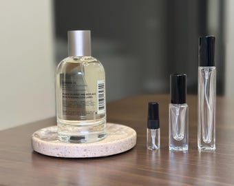Op de afbeelding: Een heldere glazen parfumfles met een zilveren dop staat op een kleine, ronde, gespikkelde stenen tray. Verschillende kleinere glazen flesjes en spuitflessen zijn in de buurt gerangschikt. Op het etiket van de parfumfles staat "LAVANDE 31".