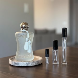 May include: A frosted glass perfume bottle with a silver cap and white tassels, displayed on a stone tray. Three smaller glass spray bottles with black caps are arranged to the right. The bottles are on a dark brown wooden surface.