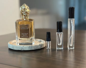 Op de afbeelding: Een grote glazen parfumfles met een gouden dop en een etiket met de tekst Maison Mataha Printemps Blanc, vergezeld door drie kleinere doorzichtige glazen verstuivers met zwarte doppen, opgesteld in een rij op een rond gespikkeld stenen dienblad.