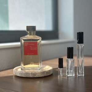May include: A rectangular perfume bottle with a red label and gold accents sits on a round stone coaster. Several smaller perfume bottles of varying sizes are arranged nearby. The bottles are clear, and the caps are black.