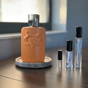 May include: An orange perfume bottle with a silver cap and a marble coaster. The bottle has a crest design and the words "Parfums de Marly". Several smaller perfume vials are next to the bottle.