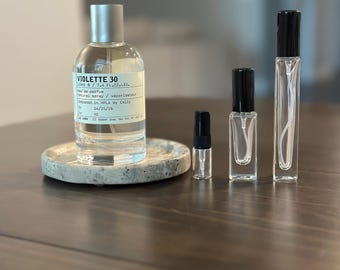 Op de afbeelding: Een grote, doorzichtige glazen parfumfles met het label VIOLETTE 30 en een zilveren dop op een gespikkelde stenen onderzetter, vergezeld door drie kleinere, doorzichtige glazen verstuivers van verschillende hoogtes met een zwarte dop.