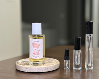 Op de afbeelding: Een verzameling parfumflesjes van verschillende formaten. De grootste fles, met een witte dop, staat op een ronde, gespikkelde stenen onderzetter. Het etiket luidt "Malaia" en "Mellow Moments". Andere flessen zijn transparant met zwarte sproeikoppen.