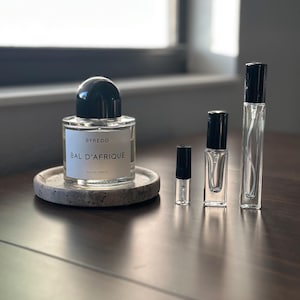 May include: A clear glass perfume bottle with a black cap and the text "BYREDO BAL D'AFRIQUE" sits on a small, round, stone coaster. Several smaller perfume vials with black caps are next to the bottle. The bottles are on a dark wooden surface.
