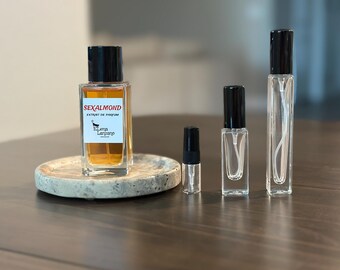 Op de afbeelding: Een rechthoekige glazen parfumfles met een zwarte dop en een wit etiket met de tekst SEXALMOND EXTRAIT DE PARFUM Elena Lanzano, vergezeld door drie kleinere doorzichtige glazen verstuivers van verschillende hoogtes met zwarte spuitdoppen.