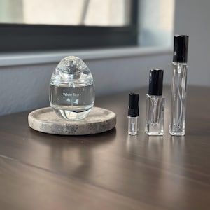 May include: A collection of clear glass perfume bottles and vials. The largest bottle is oval-shaped and sits on a small, round stone tray. The text "White Rice" is printed on the bottle. Several smaller vials with black caps are arranged to the right.