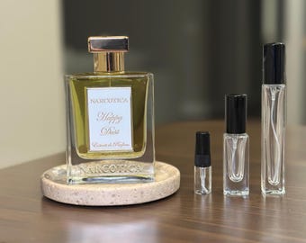 Op de afbeelding: Een parfumfles met een gouden dop en de tekst "NARCOTICA Happy Dust" staat op een kleine, ronde, beige stenen tray. Verschillende kleinere parfumflesjes van verschillende formaten zijn rechts van de fles gerangschikt. De flessen staan op een donkere houten ondergrond.