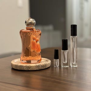 May include: An ornate, copper-colored perfume bottle with a silver cap and orange tassel sits on a marble coaster. Several clear, empty perfume vials of varying sizes stand nearby, ready for filling. The items are displayed on a dark wooden surface.