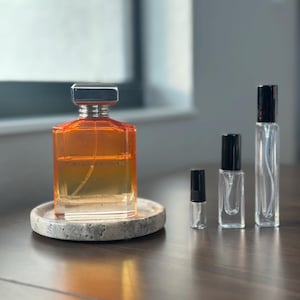 May include: An orange-tinted perfume bottle with a silver cap sits on a small, round, stone coaster. Several smaller, clear glass perfume bottles with black caps are arranged to the right. The bottles are on a dark wooden surface.