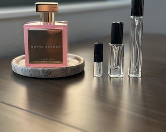Op de afbeelding: Een roze parfumfles met een gouden dop en de tekst "Guava Sorbet" op de voorkant, samen met verschillende kleine glazen parfummonsterflesjes. De parfumfles staat op een ronde, grijze stenen onderzetter.