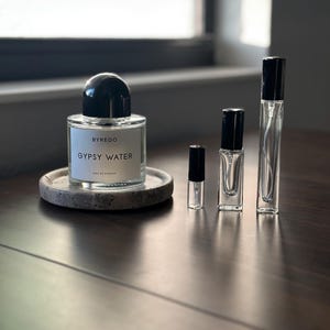 May include: A collection of Byredo Gypsy Water perfume bottles and vials. The main bottle is a clear, rectangular shape with a black cap, resting on a small, round stone coaster. Several smaller vials of varying sizes are arranged beside it, all with black caps.