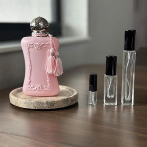 May include: A pink perfume bottle with a silver cap and pink tassels sits on a round, stone-like tray. Several small, clear spray bottles with black caps are arranged to the right. The background is a blurred interior.