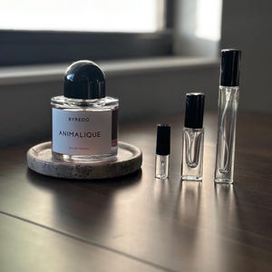 May include: A perfume bottle and several smaller vials are arranged on a wooden surface. The main bottle, labeled "BYREDO ANIMALIQUE EAU DE PARFUM," has a black cap and sits on a round, speckled coaster. The smaller vials have black caps.