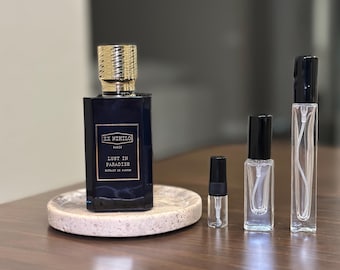 Op de afbeelding: Een donkerblauwe parfumfles met een gouden dop en de tekst "EX NIHILO PARIS LUST IN PARADISE" staat op een kleine, ronde, gespikkelde stenen tray. Er staan ​​verschillende kleinere, heldere spuitflessen naast.