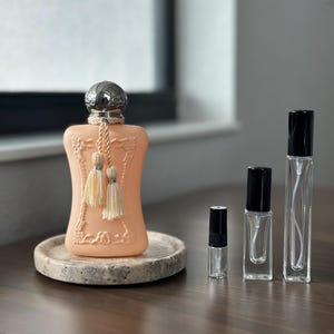 May include: A peach-colored perfume bottle with a silver cap and tassel detail, displayed on a stone tray. Several clear glass perfume sample bottles with black caps are arranged nearby, showcasing a fragrance collection.