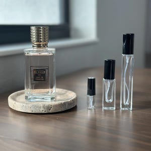 May include: A clear perfume bottle with a gold cap and the text "EX NIHILO LUST IN PARADISE" sits on a small, round, stone tray. Three smaller, clear spray bottles with black caps are next to it. The bottles are on a dark wood surface.