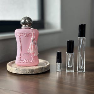 May include: A pink perfume bottle with a silver cap and tassel sits on a marble tray. The bottle is labeled "DELINA EXCLUSIF." Several small, clear glass perfume vials with black caps are arranged nearby on a wooden surface.