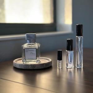 May include: A collection of perfume bottles of varying sizes, including a large square bottle with a silver cap and a smaller sample vial. The bottles are arranged on a dark wooden surface, with a round stone coaster. The text "Maison Francis Kurkdjian Paris L'Homme &Agrave; la rose" is visible.