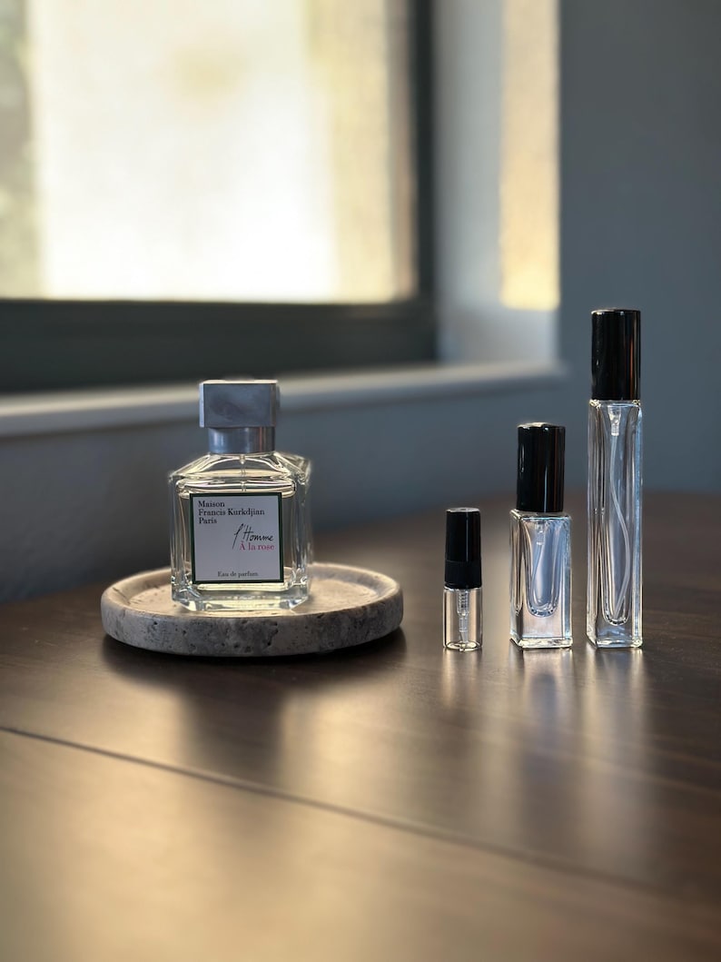 May include: A collection of perfume bottles of varying sizes, including a large square bottle with a silver cap and a smaller sample vial. The bottles are arranged on a dark wooden surface, with a round stone coaster. The text "Maison Francis Kurkdjian Paris L'Homme &Agrave; la rose" is visible.