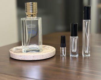 Op de afbeelding: Een verzameling heldere glazen parfumflesjes van verschillende formaten. De grootste fles heeft een goudkleurige dop en staat op een ronde, gespikkelde, beige stenen tray. De kleinere flesjes hebben zwarte doppen. Op een donkerbruin houten oppervlak.