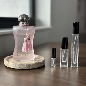 May include: A collection of perfume bottles and samples. The largest bottle is a light pink color with a silver cap and pink tassel. Several smaller clear glass bottles with black caps are also present, arranged on a wooden surface.