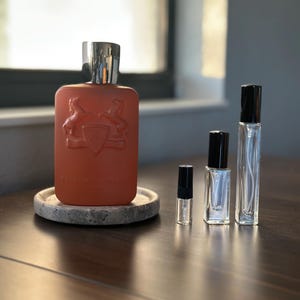 May include: An orange-red perfume bottle with a silver cap, embossed with a crest, sits on a small, round stone coaster. Several smaller glass perfume vials with black caps are arranged nearby, suggesting a fragrance sample set.