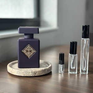 May include: A dark purple perfume bottle with a gold emblem sits on a small, round, stone tray. Several clear glass perfume sample bottles with black caps are arranged to the right of the perfume bottle. The background is blurred.