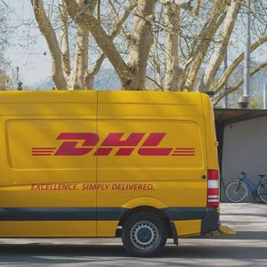 May include: A yellow DHL delivery van with the DHL logo in red. The van has the text "EXCELLENCE. SIMPLY DELIVERED." on the side. A person in a red and yellow vest stands near the van.
