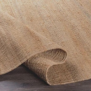 May include: A rolled-up, beige jute rug. The rug has a woven texture and a natural, earthy tone. The edge of the rug is curved, revealing the underside. The rug is on a dark wooden surface.