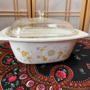 May include: A white casserole dish with a clear glass lid. The dish has a floral pattern with yellow and white flowers. The dish is on a patterned black tablecloth.