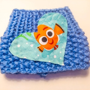 May include: A blue knitted hat with a heart-shaped patch featuring a cartoon orange fish with white eyes and a smile. The fish is on a light blue background with white dots.